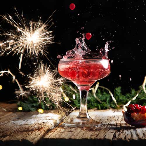 Ein Cocktailglas mit einem roten Getränk spritzt dramatisch, umgeben von Wunderkerzen und festlichen Lichterketten auf einer Holzoberfläche. Im Hintergrund ist eine Granatapfelhälfte zu sehen, die zur festlichen Atmosphäre beiträgt.