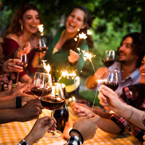 Eine Gruppe von Menschen sitzt an einem Tisch im Freien, stößt mit Weingläsern an und hält Wunderkerzen in den Händen. Sie lächeln und feiern, umgeben von Grün in einer festlichen Atmosphäre.