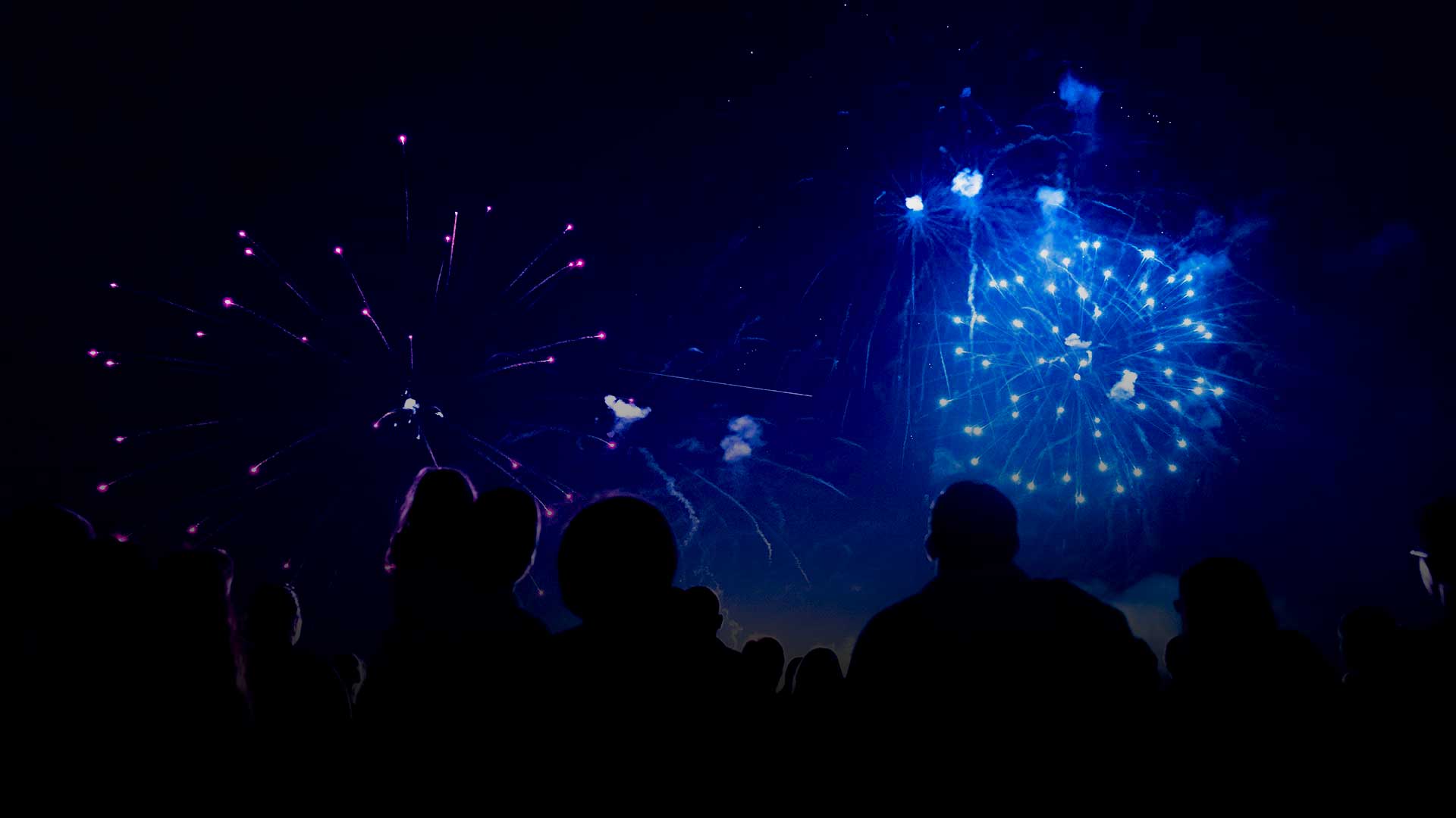Silhouetten einer Menschenmenge, die zusieht, wie leuchtend blaue und violette Feuerwerke den Nachthimmel erhellen.