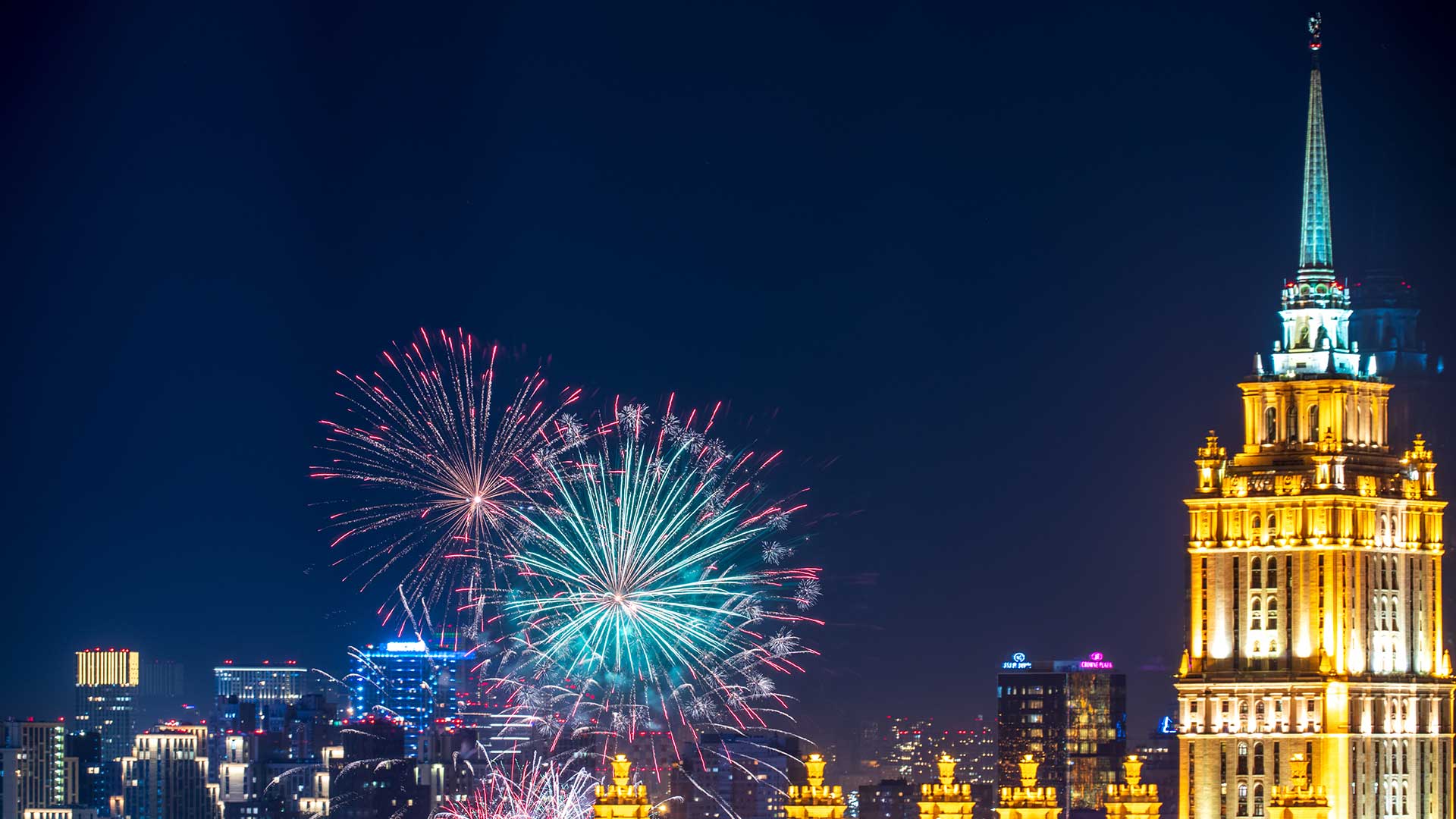 Nächtliche Stadtlandschaft mit leuchtendem Feuerwerk, das den Himmel erhellt. Auf der rechten Seite ist ein hohes, beleuchtetes Gebäude zu sehen, während die Lichter der Stadt und die Wolkenkratzer eine geschäftige urbane Kulisse bilden.